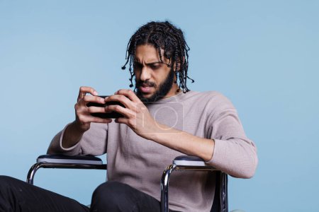 Photo for Concentrated man in wheelchair using application on mobile phone with tense facial expression. Person with physical impairment playing difficult online game on smartphone - Royalty Free Image