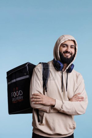 Foto de Hombre árabe sonriente posando con la mochila de servicio de entrega de alimentos y mirando a la cámara con expresión alegre. Repartidor feliz de pie con bolsa termal con retrato de almuerzo restaurante - Imagen libre de derechos