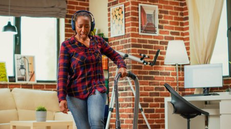 Téléchargez les photos : African american woman listening to music on headset and vacuuming floors in living room, using vacuum cleaner and having fun spring cleaning. Modern adult dancing and singing. Handheld shot. - en image libre de droit