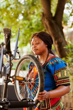 Foto de La joven ciclista activa enfocada asegura que la bicicleta esté bien preparada para aventuras al aire libre asegurando diligentemente el caucho de los neumáticos. saludable y deportivo afroamericano dama agarrando rueda de bicicleta para - Imagen libre de derechos