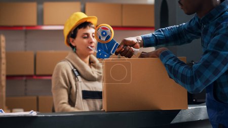 Photo for Young depot worker putting adhesive tape on packages, preparing order with merchandise in warehouse space. Male supervisor sealing off cardboard boxes with goods, delivery service. - Royalty Free Image