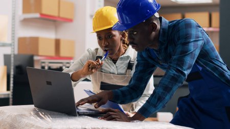 Photo for African american employees reading products on laptop and looking at goods packed in cardboard boxes in depot. Warehouse manager and worker checking stock logistics, small business. Handheld shot. - Royalty Free Image