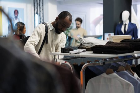 Photo for African american man browsing rack with casual clothes while shopping in mall fashion department. Boutique store customer examining menswear apparel while choosing trendy outfit - Royalty Free Image