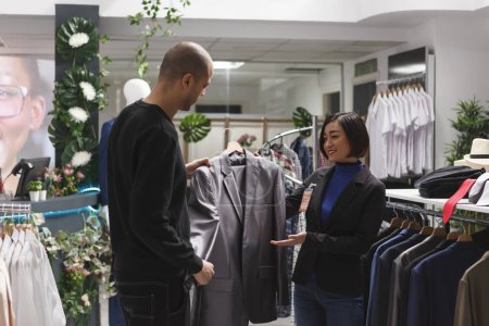 Photo for Shopping mall boutique smiling asian woman seller assisting arab man client in selecting jacket. Clothing store worker holding formal apparel hanger, showing outfit to customer, giving fashion advice - Royalty Free Image
