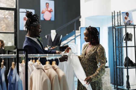 Foto de Mujer afroamericana comprando ropa para el embarazo en una boutique moderna, trabajadora ayudando al cliente con la camisa adecuada. Cliente embarazada comprando mercancía de moda en la tienda de ropa - Imagen libre de derechos