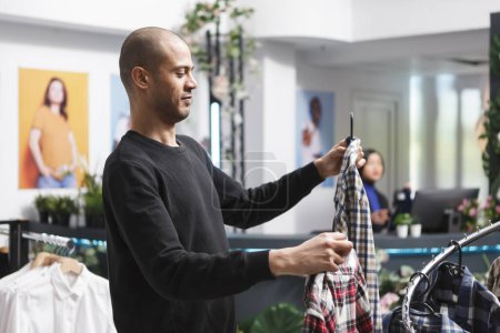 Photo for Arab man standing in mall clothing store, searching for plaid shirt and choosing among two options. Shopping center boutique buyer examining garment before making purchase - Royalty Free Image