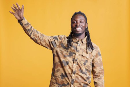 Photo for African american man waving with palm at camera, smiling person being friendly in studio. Cheerful person doing salute gesture to greet people while standing over yellow background - Royalty Free Image