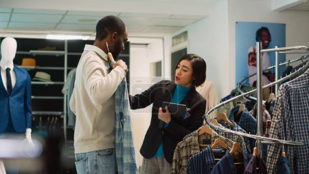 Photo for Diverse people examining clothes on racks to do shopping, woman working as store assistant in shopping center to help man choose shirts. Client looking at fashion merchandise. Handheld shot. - Royalty Free Image