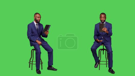 Photo for Business manager talking on online videoconference call sitting on chair in studio, full body green screen backdrop. Young entrepreneur using tablet with remote videocall meeting. - Royalty Free Image