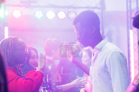 Téléchargez les photos : Couple afro-américain montrant les mains se déplace tout en improvisant bataille de danse dans une boîte de nuit sombre éclairée par des lumières. Jeunes danseurs faisant la fête à la discothèque dans un club bondé - en image libre de droit