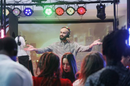 Man dj in headphones performing on nightclub stage at electronic music festival while people dancing. Young musician mixing tracks while clubbers crowd having fun on dancefloor