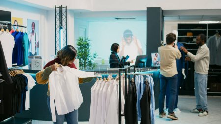 Photo for Female customer looking at merchandise in clothing store, shopping for new trendy collection. Young woman checking clothes hanging in department store, modern fashion boutique. - Royalty Free Image