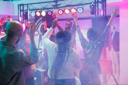 Téléchargez les photos : Foule énergique dansant et profitant de la discothèque sur piste de danse avec des projecteurs colorés. Les gens sautent, s'amusent et se déplacent vers la musique bat avec les mains levées dans la boîte de nuit - en image libre de droit