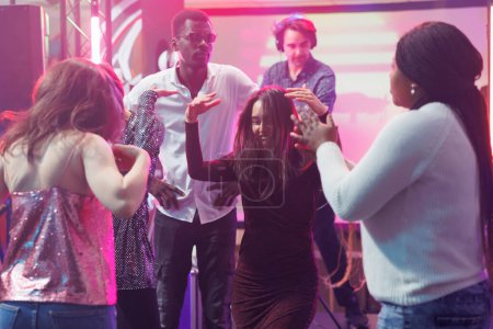 Téléchargez les photos : Clubbers insouciants amis danser et s'amuser ensemble dans une boîte de nuit. Diverses personnes faisant des mouvements énergiques au rythme de la musique sur la piste de danse au club disco rassemblement social - en image libre de droit