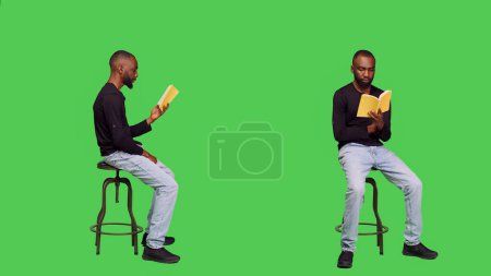 Téléchargez les photos : Une personne concentrée lisant un livre de contes en studio, assise sur une chaise sur un écran vert complet. Jeune homme optimiste profitant de roman conférence sur la caméra, activité de loisirs avec la littérature. - en image libre de droit