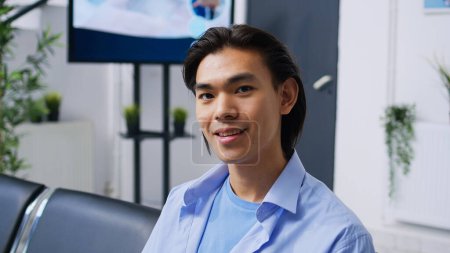 Photo for Young adult waiting to attend checkup visit with medic, sitting on chair in hospital reception. Asian patient preparing for consultation, having medical appointment. Medicine service and support - Royalty Free Image