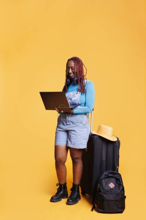 Foto de Turista femenina usando laptop en estudio, esperando salir en viaje de vacaciones. Mujer joven con bolsas de carro y equipaje que sostiene la PC, viajando al extranjero y sentado sobre fondo naranja. - Imagen libre de derechos