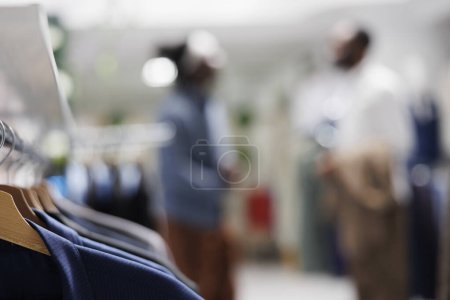 Photo for Male shirts hanging on rack in shopping mall with customer and assistant choosing clothes on blurred background. Menswear on hangers in fashion boutique close up selective focus - Royalty Free Image