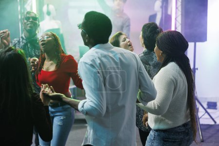 Téléchargez les photos : Diverses personnes clubbing et danser ensemble à l'événement de fête discothèque dans la boîte de nuit. Jeunes amis se tenant la main, chantant et se relaxant sur la piste de danse bondée au rassemblement social - en image libre de droit