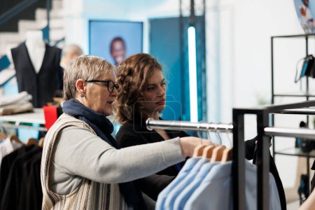 Téléchargez les photos : Employé du showroom aider les femmes âgées à choisir des vêtements à la mode, discuter de tissu dans la boutique moderne. Achat de vêtements décontractés, achat de marchandises à la mode dans un magasin de vêtements - en image libre de droit