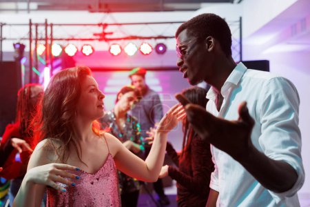 Photo for Man and woman diverse friends dancing together and chatting at nightclub disco party. Young multiracial couple clubbing and having fun on dancefloor at discotheque in club - Royalty Free Image