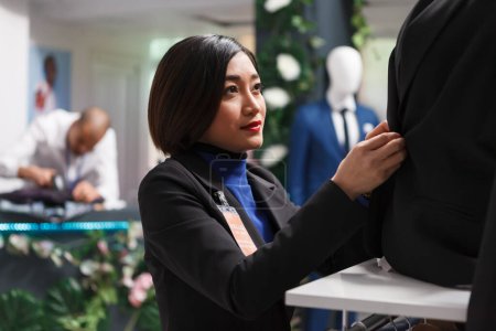 Photo for Clothing store asian woman seller showcasing stylish outfit on mannequin while fastening jacket buttons. Retail center boutique worker adjusting apparel while organizing merchandise - Royalty Free Image