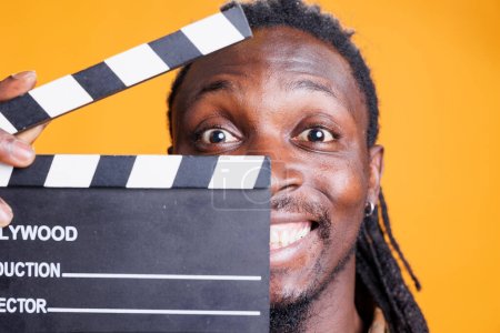 Foto de Retrato del hombre sosteniendo pizarra para cortar escenas en la industria cinematográfica, posando sobre fondo amarillo. Adultos jóvenes afroamericanos trabajando en la producción cinematográfica y cinematográfica - Imagen libre de derechos