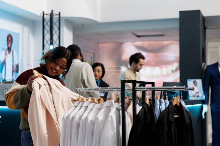 Photo for African american woman holding outfit to body, checking size and style before making purchase in boutique. Customer examining formal shirt fit on hanger in clothing store - Royalty Free Image