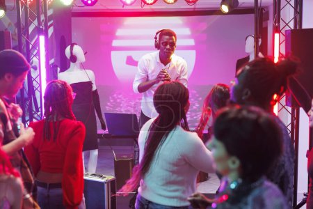 Foto de Dj mezclando música y cantando en el micrófono mientras la multitud baila y anima en la discoteca. Músico afroamericano actuando en el escenario y hablando con el público en el club - Imagen libre de derechos