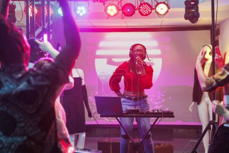 Musician singing and playing electronic music on stage in nightclub. African american woman dj using microphone and sound controller equipment while performing in crowded club
