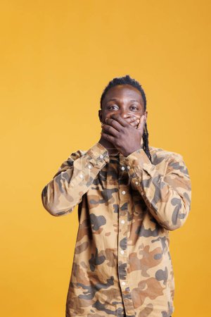 Photo for Young adult covering mouth with palms, doing doing three wise monkeys symbolic sign, standing in studio over yellow background. African american man showing silence gesture, not speaking concept - Royalty Free Image