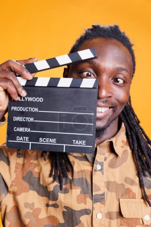 Téléchargez les photos : Jeune adulte tenant l'ardoise filmographique, posant confiant devant la caméra en studio sur fond jaune. Homme afro-américain travaillant dans l'industrie de la production cinématographique - en image libre de droit