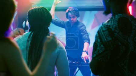 Photo for Group of friends dancing in the club, enjoying live performance show with DJ mixing station on nightclub stage. Young people partying together on dance floor with disco lights. Tripod shot. - Royalty Free Image