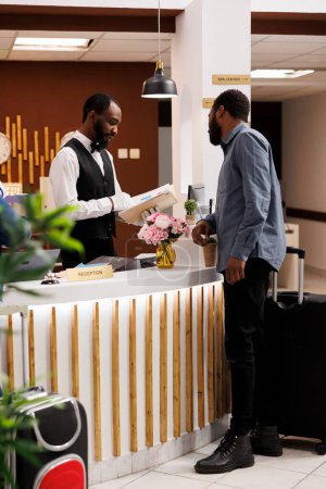 Téléchargez les photos : Jeune agent afro-américain à la réception confirmant la réservation. Réceptionniste masculin debout derrière le comptoir de réception parlant avec le touriste arrivant à l'hôtel, vérifiant la disponibilité des chambres - en image libre de droit