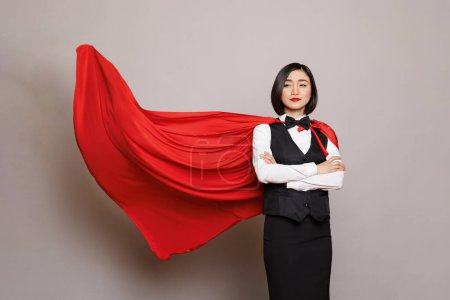 Photo for Confident asian waitress standing with folded arms and posing in fluttering superman red cape. Young attractive woman receptionist dressed in hero cloak and catering service uniform - Royalty Free Image
