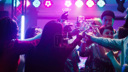 Happy diverse people clinking glasses at disco party, drinking alcohol and saying cheers on dance floor. Men and women partying and having fun at nightclub, raising drinks at event.