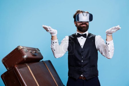 Photo for Modern doorkeeper trying on vr headset against blue background, using virtual reality technology in studio. Young man with bellhop occupation standing next to luggage, tourism concept. - Royalty Free Image