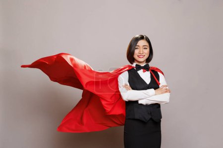 Photo for Attractive young woman wearing waitress uniform and fluttering superman cloak while posing in studio portrait. Confident restaurant employee dressed in superhero red cape - Royalty Free Image