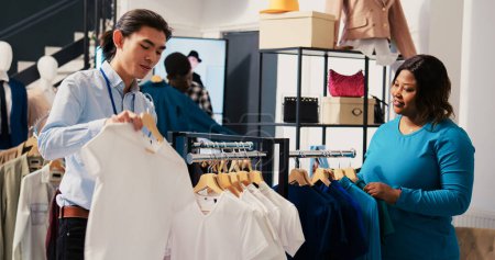 Foto de Shooper pidiendo ayuda a los trabajadores asiáticos con camisas de moda, discutiendo el material de mercancías en la boutique moderna. Cliente afroamericano comprando ropa elegante en tienda de ropa - Imagen libre de derechos