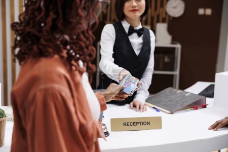 Photo for Young hotel guest paying for reservation at reception counter, using card at pos terminal. Front desk staff welcoming people at modern luxury resort, electronic payment at check in. - Royalty Free Image