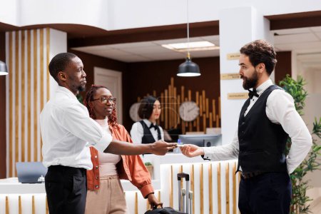 Photo for Bellboy accepting cash from hotel guest in reception lobby, offering luxury services to carry luggage at front desk. Young man giving tip to hotel concierge, helping with suitcases. - Royalty Free Image
