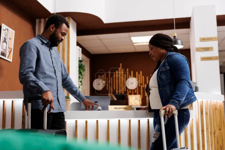 Photo for Young African American man tourist ringing service bell on hotel reception desk, waiting for check-in. Black couple with suitcases wait for receptionist to help with registration process - Royalty Free Image