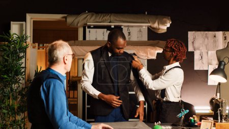 Young customer trying on suit jacket in atelier, making sure it fits with the right measurements. Experienced tailor and assistant designing custom made blazer, fitting in workshop.
