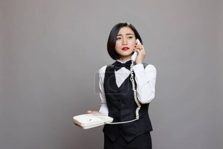 Photo for Young confident asian waitress in uniform talking on landline phone and looking away. Serious woman receptionist holding retro telephone and speaking with restaurant staff - Royalty Free Image