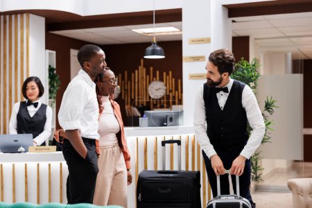 Photo for Hotel steward carrying bags at resort, offering to help guests with their luggage at reception desk in lobby. Bellboy providing excellent luxury service to tourists, trolley bags. - Royalty Free Image