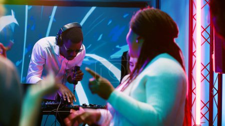 African american DJ having fun at club, mixing electronic music on audio station at discotheque. Young man feeling happy partying with people on stage, using sound panel at nightclub.