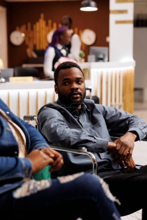 Photo for Young African American man tourist on wheelchair arriving at hotel for disabled, sitting in lobby talking with girlfriend or wife while waiting for check-in procedure. Using wheelchair during travel - Royalty Free Image