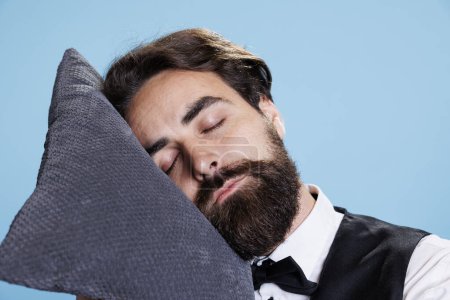 Photo for Classy bellboy falling asleep on pillow while he stands in studio, feeling extremely worn out and exhausted. Professional employee doorman taking a quick nap on camera, expressing burnout. - Royalty Free Image