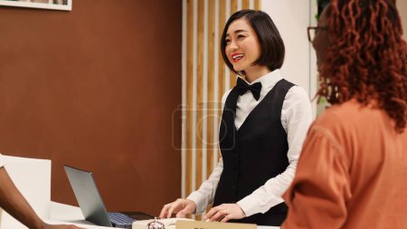 Photo for Happy friendly asian receptionist helping guests during check in process, imputing their personal information on laptop. Tourists booking in their stay in luxury hotel, assisted by hotel staff - Royalty Free Image
