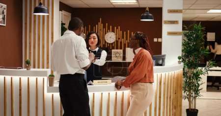 Photo for Friendly smiling asian receptionist handing contract on tablet for african american tourists to sign after upgrading room amenities. Happy relaxed guests on vacation trip checking in stylish hotel - Royalty Free Image